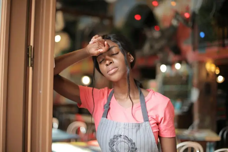 A tired woman in an apron stands in a doorway with her eyes closed, looking exhausted.